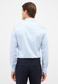 Camicia a maniche lunghe azzurra con una texture morbida, colletto alla francese e polsini con bottoni, abbinata a pantaloni scuri con tasche posteriori.