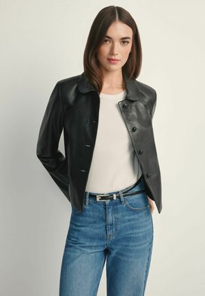 Czarna skórzana kurtka z kołnierzem, przednimi guzikami i długimi rękawami. W zestawie z białą koszulą i niebieskimi, wysokim stanem jeansami.