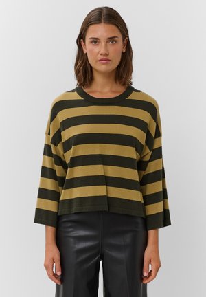 Femme portant un pull ample à larges rayures olive et moutarde avec des manches trois-quarts et un pantalon en cuir noir, debout devant un fond blanc.