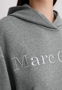 Šedá mikina s kapucí, na které je vyšitý nápis "Marc O'" ve světle šedé barvě. Vyznačuje se měkkou texturou a jednoduchým designem, zaměřuje se na pohodlí a volnočasové nošení.