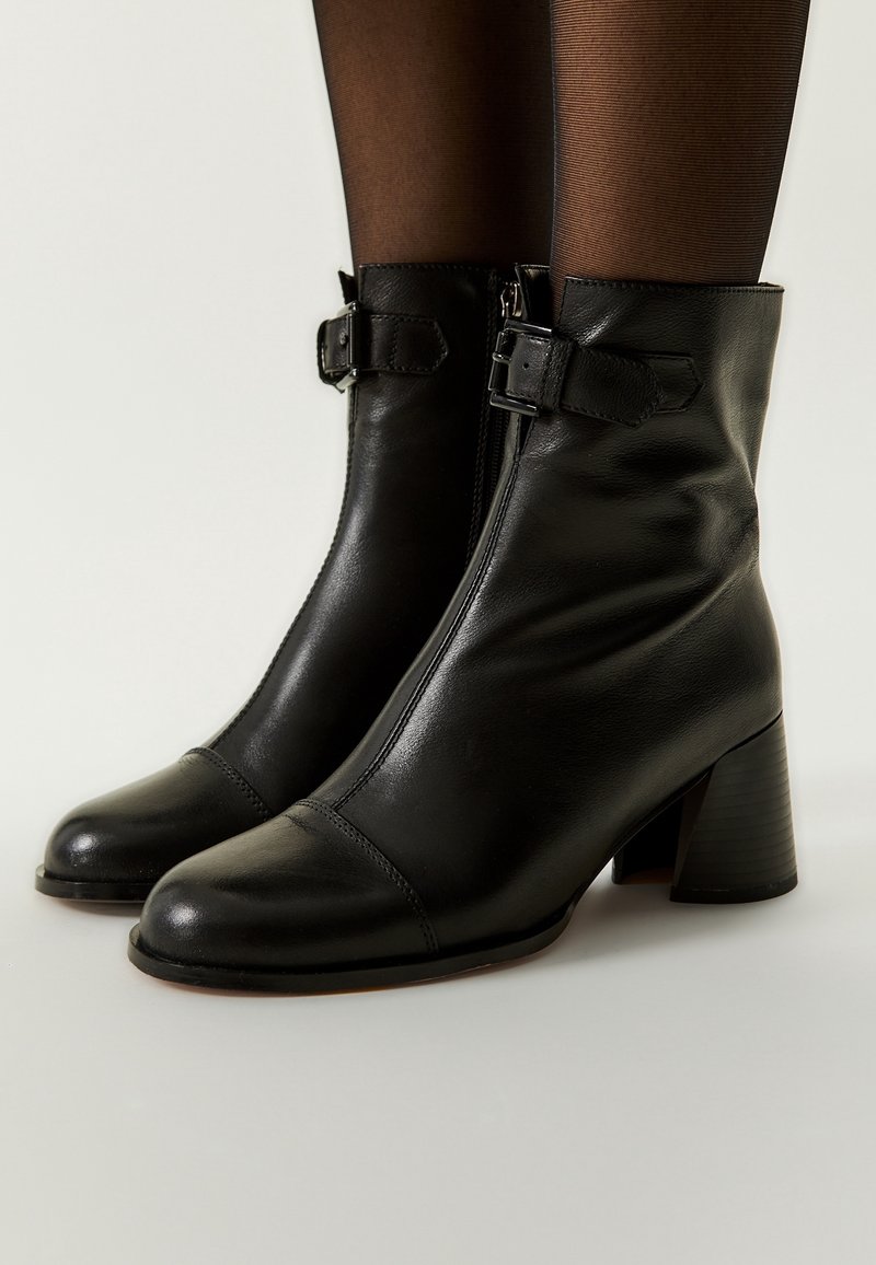 Bottines en cuir noir avec fermeture éclair latérale, bride à boucle, talons blocs bas, portées avec des collants noirs transparents sur un fond clair.