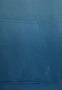 Modrá mikina z látky s žebrovanou texturou. Má přední kapsu a viditelné švy podél švů. Měkký, ležérní design.