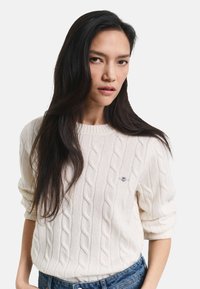 Maglione crema a trecce con colletto rotondo, caratterizzato da maniche arrotolate e un piccolo logo sul lato sinistro. Indossato con jeans blu.