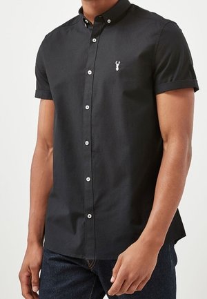Mand iført sort kortærmet skjorte med knapper og lille hvid hummerlogo på brystet og mørkeblå jeans mod ensfarvet baggrund.