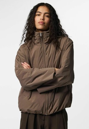 Jeune femme aux longs cheveux bouclés porte une doudoune marron, debout les bras croisés devant un fond clair uni.