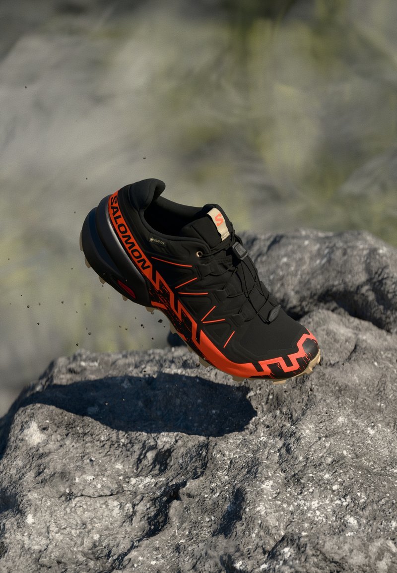 Scarpa da trail running nera e arancione con tomaia texturizzata e suola robusta, con un branding prominente e un design geometrico sui lati.