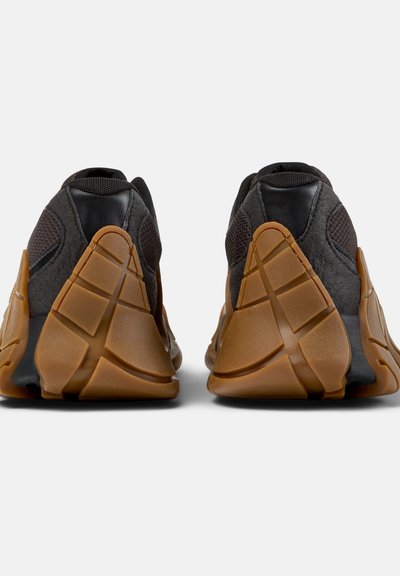 Paire de baskets noires et gris foncé avec des semelles épaisses en caoutchouc brun texturé, vues de dos sur un fond blanc.