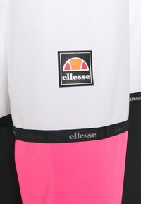 Ellesse-märkt kläder med en vit övre sektion, rosa nedre sektion, svarta accenter och en texturerad logotyp-patch i mitten.
