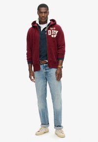 Felpa con zip color marrone bordeaux con logo, polo blu navy, jeans chiari strappati, e sneakers beige. Presenta tasche e una vestibilità casual.