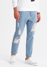 Jeans de mezclilla decolorados en azul claro con grandes desgastes y bordes deshilachados, combinados con zapatillas blancas con acentos grises.
