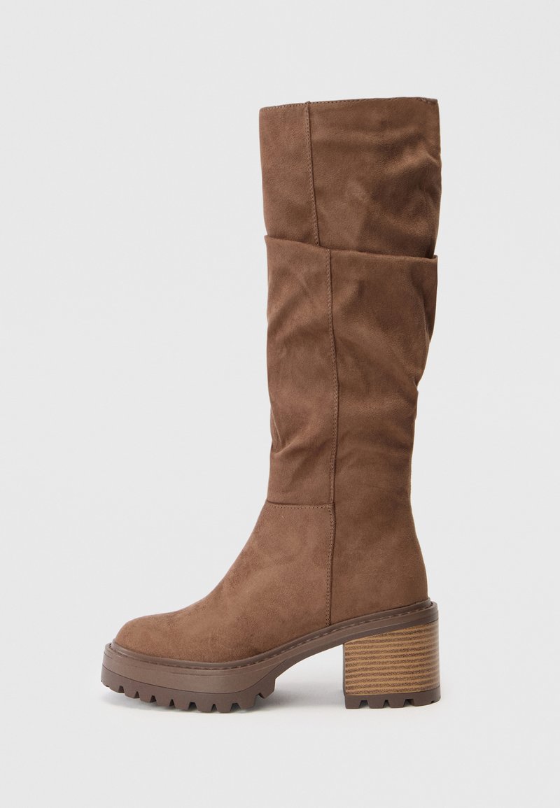 Brown suede knee-high boot with a slouchy design, a chunky striped heel, and a textured sole featuring deep grooves for traction.