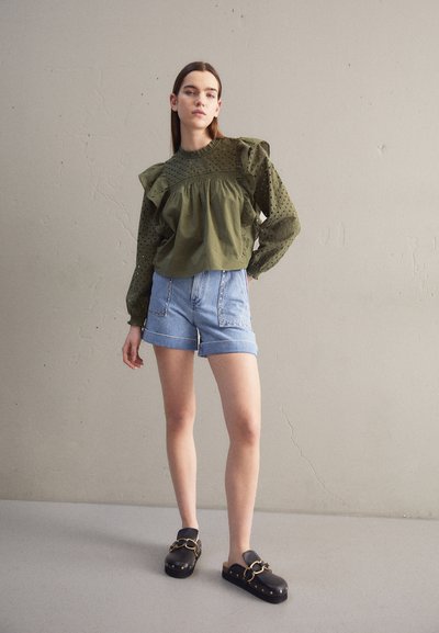 Blouse vert olive à volants avec des détails en œillets, associée à un short en denim. Sabots noirs avec des accents en chaîne dorée. Fond neutre.