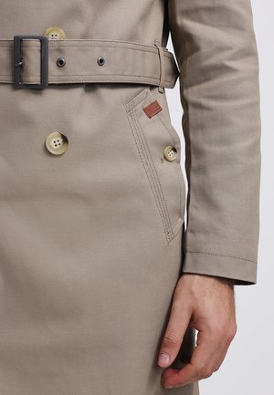 Trench-coat beige avec de gros boutons, une ceinture à boucle métallique, une poche latérale munie d'un bouton et une main visible à côté de la manche.