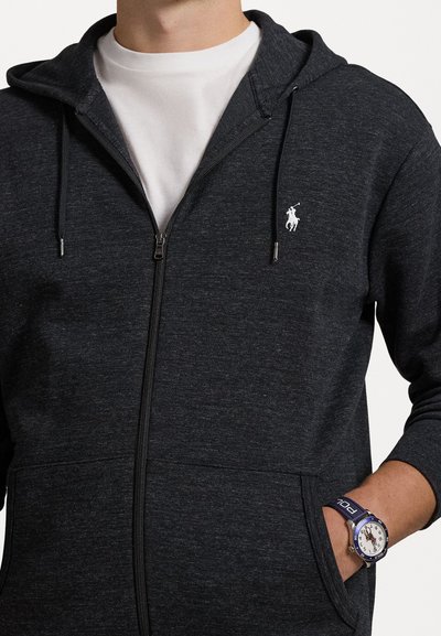 Sweatshirt cinzento com fecho, em tecido suave, apresentando um pequeno logótipo branco no peito, bolsos frontais e capuz com cordão ajustável.