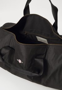 Svart stoff duffelbag med glidelåsåpning, indre lommer og topphåndtak. Har et logo på forsiden og gullglidelås.