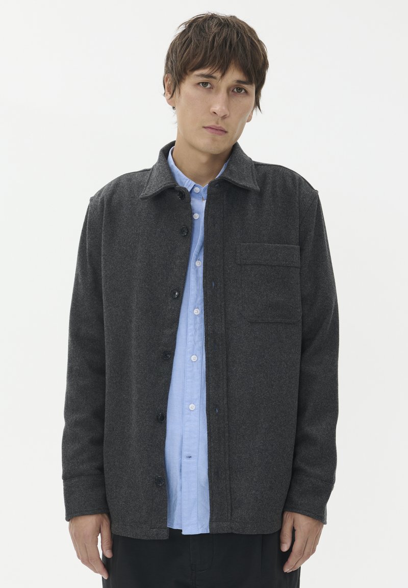 Temno siva volnena jakna s ovratnikom, zapiranjem na gumbe in žepom na prsih, oblečena preko svetlo modre srajce na gumbe.