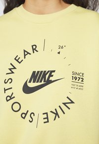 Žlutá bavlněná mikina s kulatým černým motivem obsahujícím logo "Nike", nápis "SPORTSWEAR" a souřadnice. Design s kulatým výstřihem.