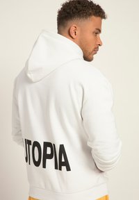 Vit hoodie gjord av mjukt tyg, med en stor svart "UTOPIA"-tryck bak, standardhuva och ribbade muddar.