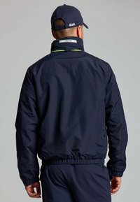 Giacca blu navale con polsini e vita elasticizzati. Presenta un colletto alto e dettagli riflettenti. La vista posteriore mostra un tessuto liscio e texture.