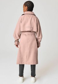 Rosa trenchcoat med en kortad baksida, långa ärmar och midjebälte. Flätat hår och gråbyxor, tillsammans med vita sneakers.