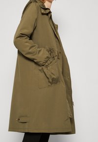 Olivgrüner wasserdichter Mantel mit Kapuze, Reißverschluss vorne, elastischen Bündchen und Fronttasche. Verfügt über goldfarbene Beschläge und eine lockere Passform.