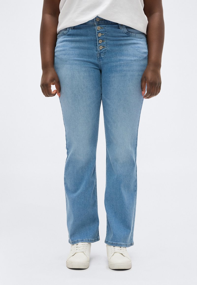 Άτομο που φοράει ανοιχτόμπλε τζιν ψηλόμεσο με πέντε μεταλλικά κουμπιά και λευκά αθλητικά παπούτσια, στέκεται μπροστά σε απλό φόντο.