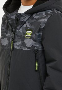 Černá bunda s šedým maskáčov�ým vzorem na horní části hrudi, přední zip, dvě kapsy na zip a táhla s logem "THREADBOYS".