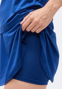 Χέρι σηκώνει μπλε φούστα για τρέξιμο αποκαλύπτοντας εφαρμοστά συμπιεστικά σορτς κάτω από αυτήν στον γυμνό μηρό.