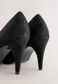 Paire d'escarpins en daim noir à talons hauts avec bouts pointus, vus de dos sur un fond clair et neutre.