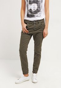 Persona con pantalones verde oliva ajustados y un cierre de cuatro botones, camiseta gráfica blanca y zapatillas blancas, de pie con las manos cerca de los bolsillos.