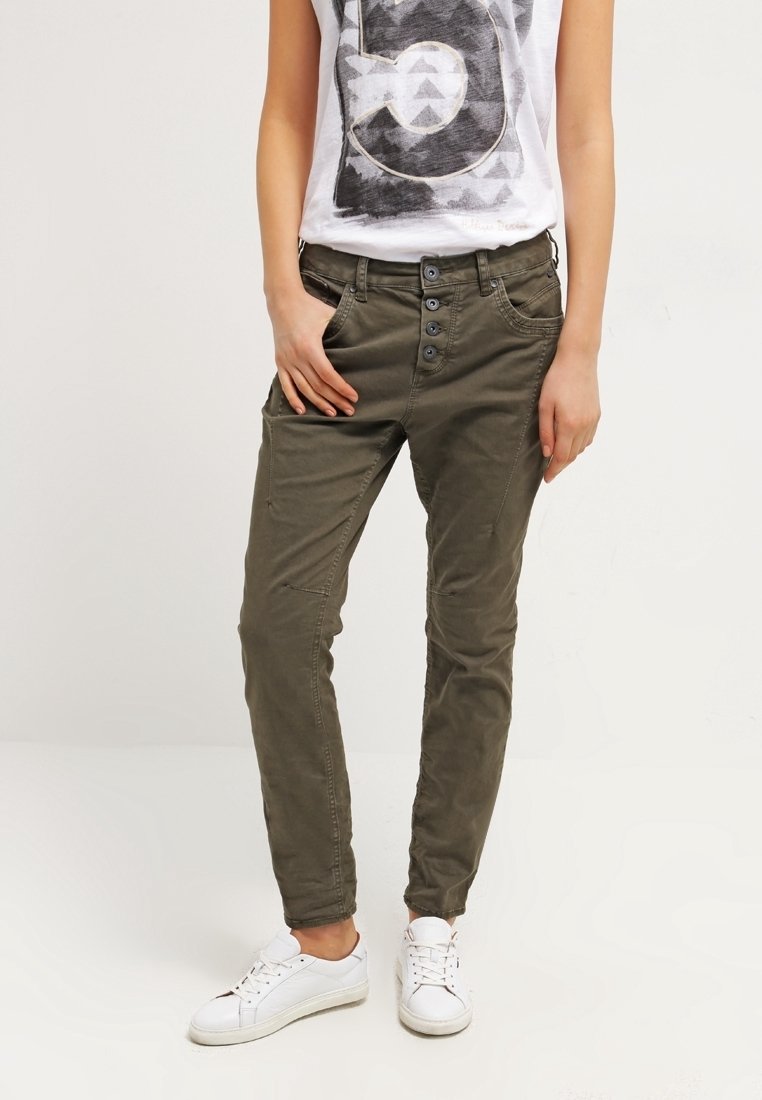 Persona con pantalones verde oliva ajustados y un cierre de cuatro botones, camiseta gráfica blanca y zapatillas blancas, de pie con las manos cerca de los bolsillos.