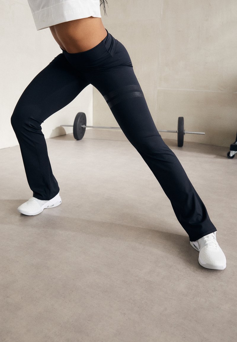 Schwarze sportliche Leggings mit einem enganliegenden Bund und dezenten Mesh-Details, kombiniert mit weißen Sportschuhen auf einem neutralen Boden.