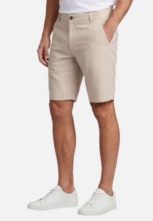 Man wearing beige knee-length shorts, white sneakers, and white shirt, standing with one hand in pocket against a plain background.