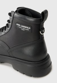 Botine negre din piele cu finisaj mat, având orificii argintii, o talpă din cauciuc texturată și un etichetă de marcă pe lateral.