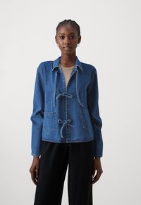 Chaqueta de mezclilla en azul medio con mangas largas, un cuello y un cierre con lazo en la parte delantera. Presenta dos bolsillos frontales y un corte relajado.