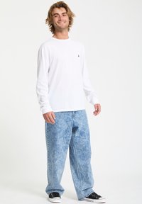 Langärmliges weißes Hemd kombiniert mit ausgebleichten blauen Schlagjeans. Das Model trägt schwarze Sneakers. Einfaches Design mit minimalistischen Logodetails.