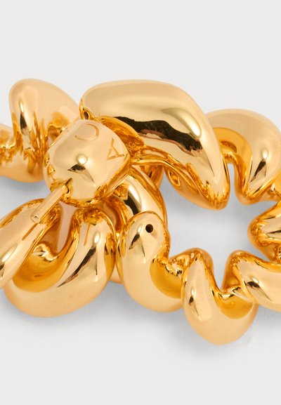 Λαμπερό βραχιόλι με χρυσούς στριφογυριστούς κρίκους αλυσίδας και γεωμετρικό κούμπωμα που φέρει χαραγμένα σύμβολα σε ανοιχτό φόντο.