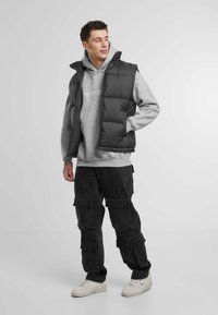 Svart polstret vest over en grå hettegenser, sammen med mørke cargo-bukser og hvite joggesko. Enkelt design med glidelås og høy krage.
