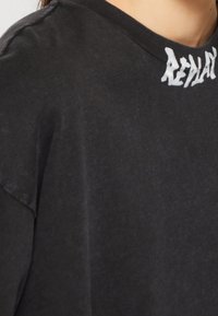 Camiseta de algodón negra con un corte holgado. Presenta un cuello redondo y letras blancas en el cuello. Textura suave con una apariencia ligeramente deslavada.