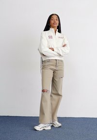 Felpa bianca con zip e logo, abbinata a jeans chiari beige strappati a zampa d'elefante e sneakers argentate. Tessuto morbido e vestibilità comoda.