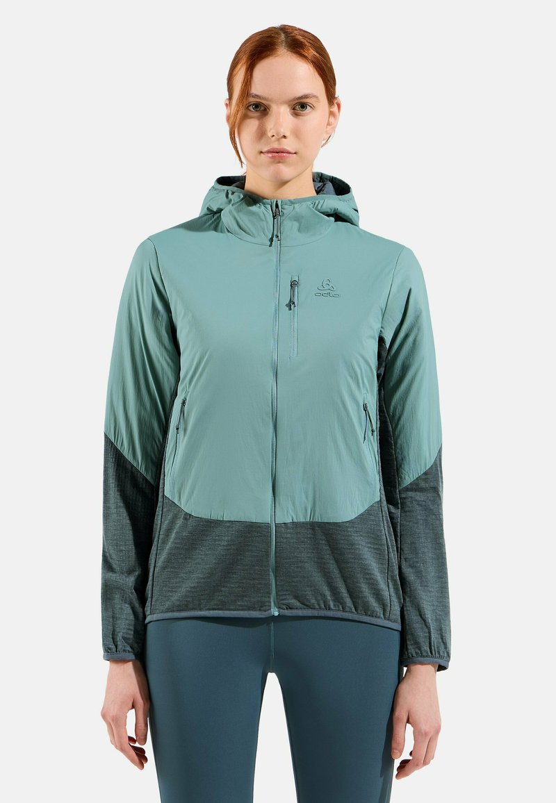 Veste légère à capuche en teal atténué avec des accents gris foncé. Dispose de poches zippées et d'une coupe ajustée pour plus de fonctionnalité.
