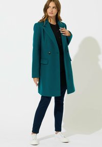 Cappotto doppiopetto color verde acqua con revers, tasche laterali e texture liscia, indossato sopra un top nero e pantaloni blu navy, abbinato a sneaker bianche.