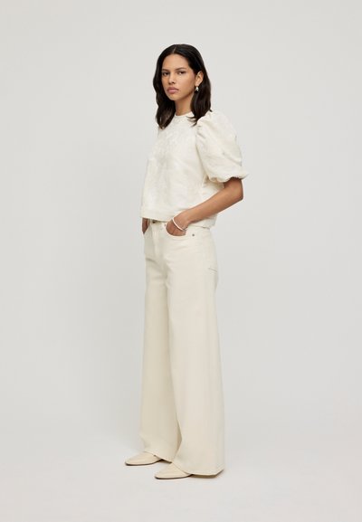 Top bianco a maniche a palloncino con una texture sottile, abbinato a pantaloni larghi color crema. La modella si presenta di profilo, mostrando accessori minimi.