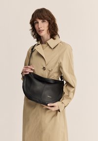 Borsa a spalla in pelle nera con forma curva, dotata di logo dorato, indossata a tracolla con tracolla regolabile. Modella in un trench beige.