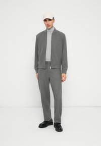 Uomo che indossa una giacca grigia con zip e pantaloni, dolcevita grigio chiaro, cappellino bianco e scarpe nere, in piedi contro uno sfondo bianco semplice.