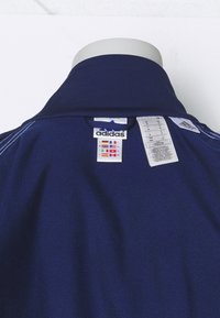 Polo shirt bleu marine avec un col côtelé, portant une étiquette logo Adidas et des étiquettes d'entretien. Le tissu semble lisse avec une légère brillance.