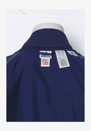 Tummansininen poolopaita, jossa on uritettu kaulus, varustettuna Adidas-logotarralla ja huoltolapulla. Kangas näyttää sileältä ja siinä on hienovarainen kiilto.