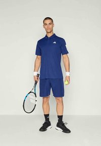 Modrá sportovní polo košile a šortky s bílými detaily. Drží tenisovou raketu a míček. Černé boty a ponožky. Jednoduchý design, sportovní střih.