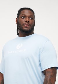 Light blue cotton t-shirt featuring white "BELSTAFF" lettering and logo, with a crew neckline and short sleeves. Smooth texture.