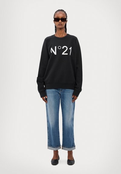 Svart hettegenser med stort hvitt "N°21"-logo, paret med lyseblå vide jeans og svarte leiligheter. Modellen har på seg store mørke solbriller.